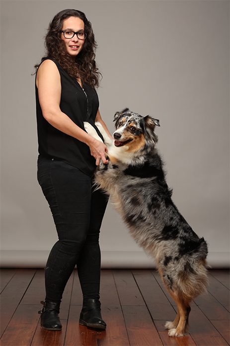 Sur la photo, on voit une femme aux cheveux bruns et bouclés, portant des lunettes et vêtue d'un haut noir sans manches et d'un pantalon noir. Elle porte également des bottines noires. À côté d'elle, se trouve une chienne de type Berger Australien, au pelage merle bleu, qui se dresse sur ses pattes arrière et pose ses pattes avant sur la femme. La chienne semble heureuse et enthousiaste. Le fond de la photo est neutre et le sol est en bois.